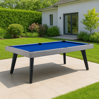 Moderne outdoor pooltafel met blauw speelvlak en zwarte schuine poten, stijlvol gepositioneerd op een licht betegeld terras in een luxueuze tuin met strak gemaaid gras, weelderige beplanting en een moderne witte woning met schuifpui op de achtergrond